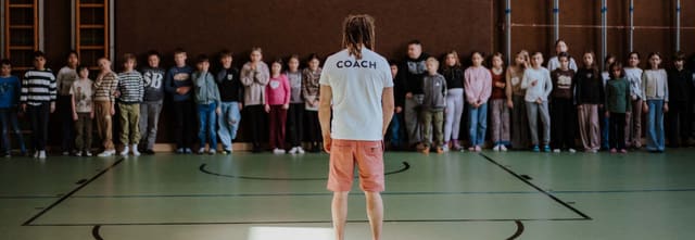 Schulklasse steht im Kreis in der Turnhalle, eine Person in der Mitte