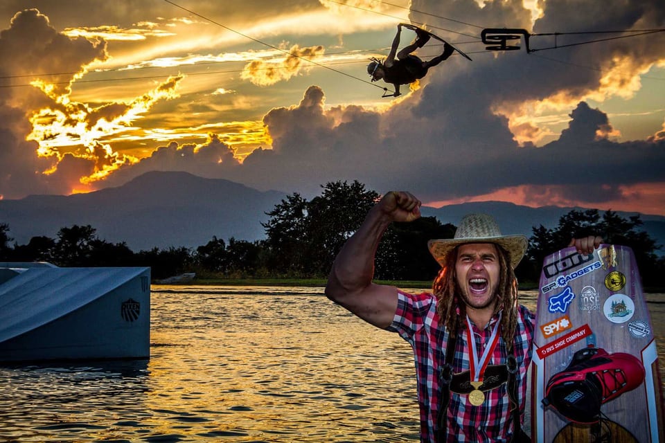Daniel Fetz jubelt mit Weltmeistermedaille und Wakeboard vor einem Sprung im Sonnenuntergang